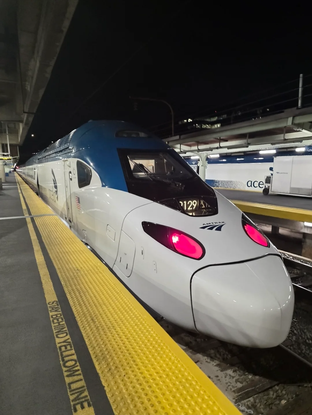 A white and blue Amtrak Acela train is stopped at a station platform at night, with its red headlights illuminated and a yellow tactile warning strip on the platform edge.