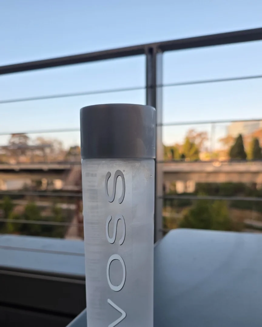 A close-up shot of a VOSS water bottle with a black cap, positioned on a balcony with a blurred background of a bridge and trees under a clear sky.
