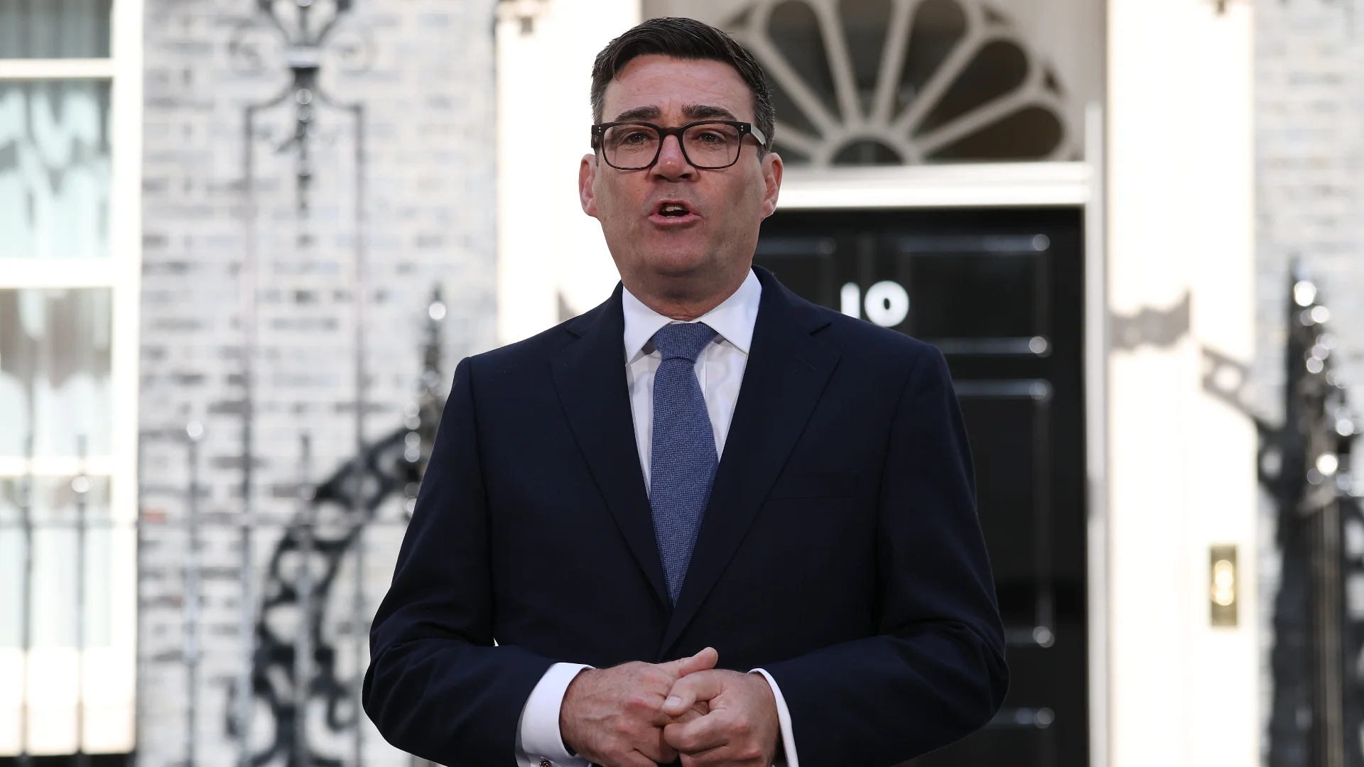 A man in a dark suit and blue tie stands in front of the black door of 10 Downing Street.